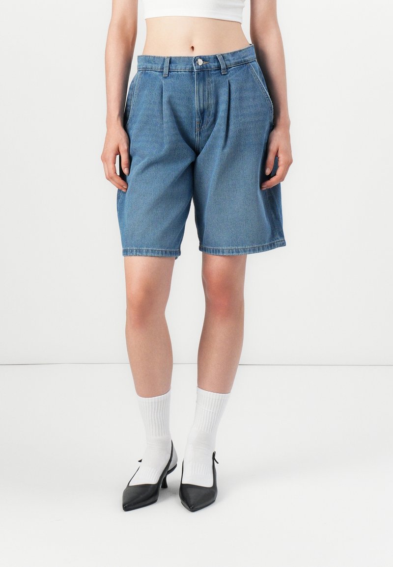Personne portant un short en jean bleu jusqu'au genou, des chaussettes blanches côtelées, des talons noirs pointus et un top court blanc, debout devant un fond uni.