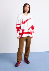 Hvid langærmet v-hals trøje med rød grafisk design og Nike-logo, parret med brune svedbukser og sort-røde sneakers på et blåt gulv.