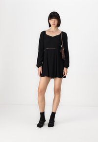 Melnas garās piedurknes kleitas ar kvadrātveida kakla izgriezumu, saplacinātu krūšu daļu un mežģīņu detaļu jostas vietā. Valkāta ar melnām zeķēm un sandalēm.