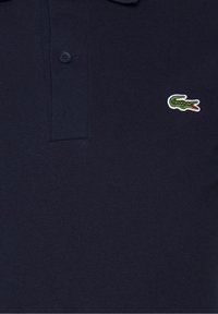 Marinblå pikétröja i texturerad bomull, med en treknapplåsning och en liten grön krokodil-logotyp på vänster bröst.