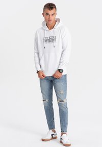 Witte hoodie met trekkoord en een voorzak, voorzien van zwarte bedrukte tekst. Gecombineerd met versleten lichtblauwe jeans en witte sneakers.