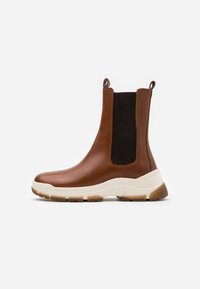 Bottes chelsea en cuir marron avec des panneaux élastiques sur les côtés, une semelle épaisse blanche et beige, et des languettes en boucle à l'arrière. Texture lisse.