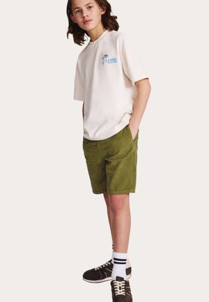 Ung person iført en off-white T-shirt med grafik, olivengrønne kordfløjls-shorts, hvide sokker med sorte striber og sorte sneakers, stående med hænderne i lommerne.