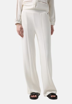 Kvinde iført hvide vidde bukser med høj talje og forreste syninger, kombineret med en cremefarvet bluse og sorte sandaler med åben tå.