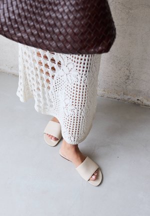 Jupe blanche en crochet avec motifs floraux, sandales beiges à enfiler et sac à main tissé foncé sur un fond de sol et mur en béton clair.