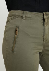 Olive green pants with a slim fit, featuring a side pocket with a gold zipper and stitching details along the waistband and seams.