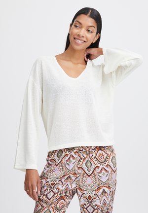 Weißer V-Ausschnitt-Pullover aus strukturiertem Stoff mit weiten Ärmeln, kombiniert mit gemusterten Hosen, die komplexe Designs in Erdtönen aufweisen.