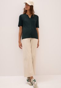 Dunkelgrüne Kurzarm-Hemdbluse, cremefarbene weit geschnittene Hose und beige Sneakers mit grünen Akzenten, vor einem hellen Hintergrund.