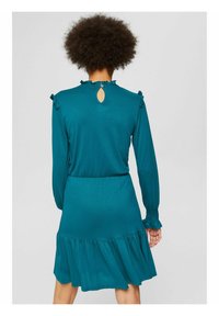 Vestido verde-água com mangas longas e detalhes franzidos nos ombros. Apresenta uma abertura em forma de olho chave nas costas e uma bainha em camadas.