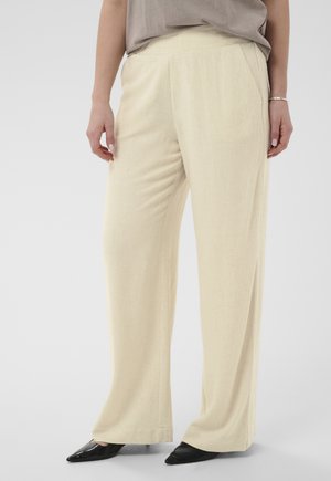 Frau trägt beige Hosen mit weitem Bein, taupefarbenes Oberteil, schwarze spitze Schuhe und ein silbernes Armband, stehend vor einem schlichten hellen Hintergrund.