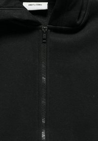 Veste à zip noire avec une texture de tissu lisse, un col haut et une fermeture éclair dissimulée à l'avant. Étiquette de la marque visible sous le col.
