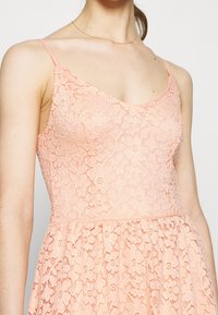 Robe en dentelle pêche avec un décolleté en V, des fines bretelles et un motif floral. Le corsage est ajusté, menant à une jupe évasée plissée.