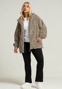 Chaqueta de forro polar texturizado gris claro con capucha, que presenta un patrón de punto trenzado y bolsillos frontales, combinada con pantalones negros y zapatillas blancas.