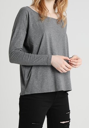 Personne portant un haut gris ample à manches longues et un jean noir déchiré, debout les mains légèrement jointes devant un fond uni.