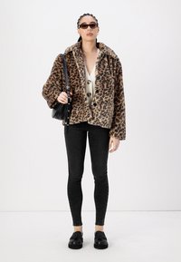 Luipaardprint nepbontjas, oversized pasvorm, zwarte knopen, gecombineerd met een lichtgekleurde top, zwarte jeans en zwarte instappers. Zwarte tas en zonnebril.