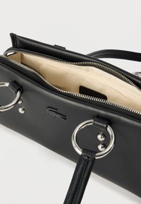 Μαύρη ορθογώνια τσάντα χειρός με ασημένιους κρίκους στα λουριά, η οποία είναι μερικώς ξεκουμπωμένη για να φαίνεται η κρεμ εσωτερική επένδυση και η μικρή εσωτερική θήκη.