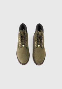 Botas de ante verde oliva con cuello de cuero negro, ojales dorados y suela de goma marrón. Presentan cordones resistentes y un borde acolchado.