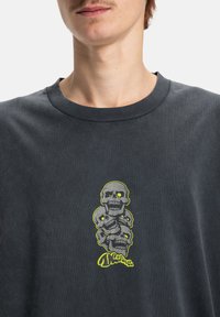 Tmavě šedé bavlněné tričko s centrální grafikou tří naskládaných lebek se žlutými akcenty a bílými detaily očí. Design s kulatým výstřihem.