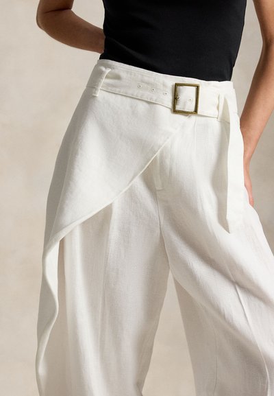Γυναίκα που φοράει λευκό παντελόνι ψηλόμεσο με φαρδιά γραμμή και ασύμμετρο ύφασμα σε συνδυασμό με ζώνη με τετράγωνη αγκράφα, συνδυασμένο με μαύρη μπλούζα.