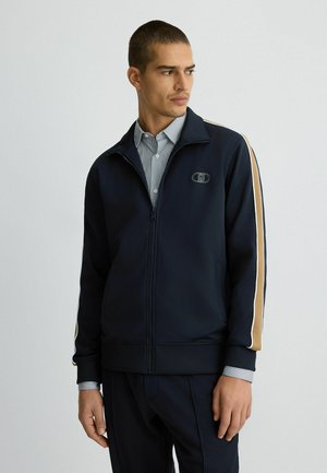 Giacca zip-up blu navy con strisce di accentuazione beige, polsini e orlo a costine, con un piccolo logo sul petto. Indossata sopra una camicia a righe con colletto.