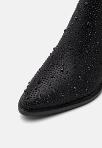 Botín negro hasta el tobillo con punta afilada, superficie texturizada con pequeños puntos elevados y cristales de rhinestone negros incrustados como detalle.