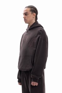 Smeđa oversized majica s kapuljačom od mekanog materijala, s prednjim džepom i suptilnim vezom. Uparena s odgovarajućim smeđim trenirkama.