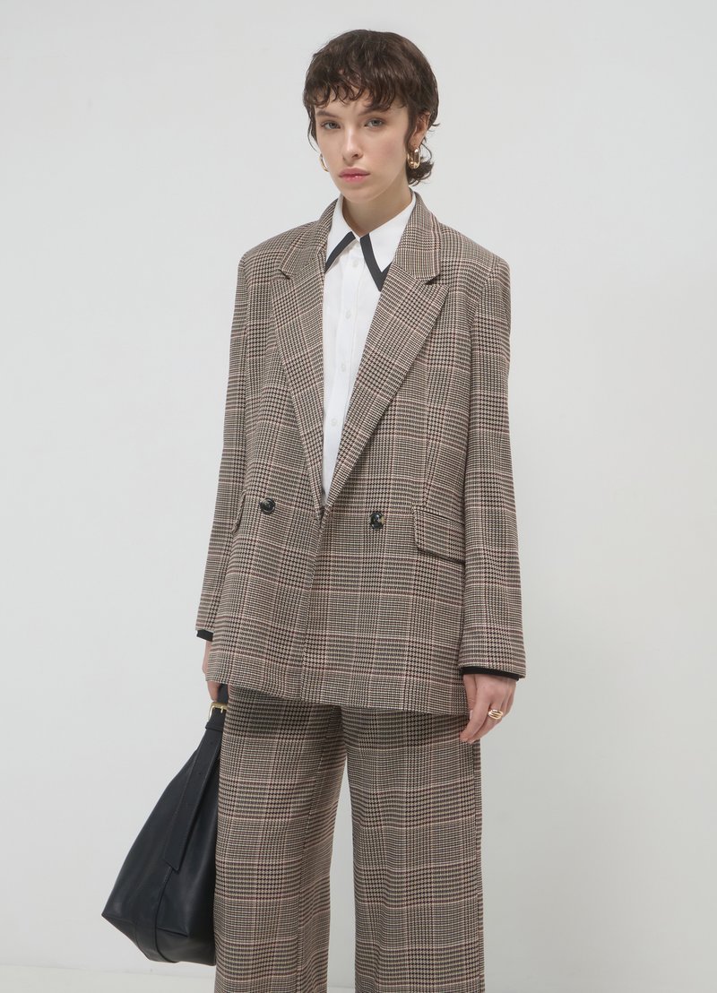 Veste oversize à double boutonnage et pantalon assorti dans un motif écossais marron, accompagnés d'une chemise blanche et d'un sac à main noir, avec une légère brillance.