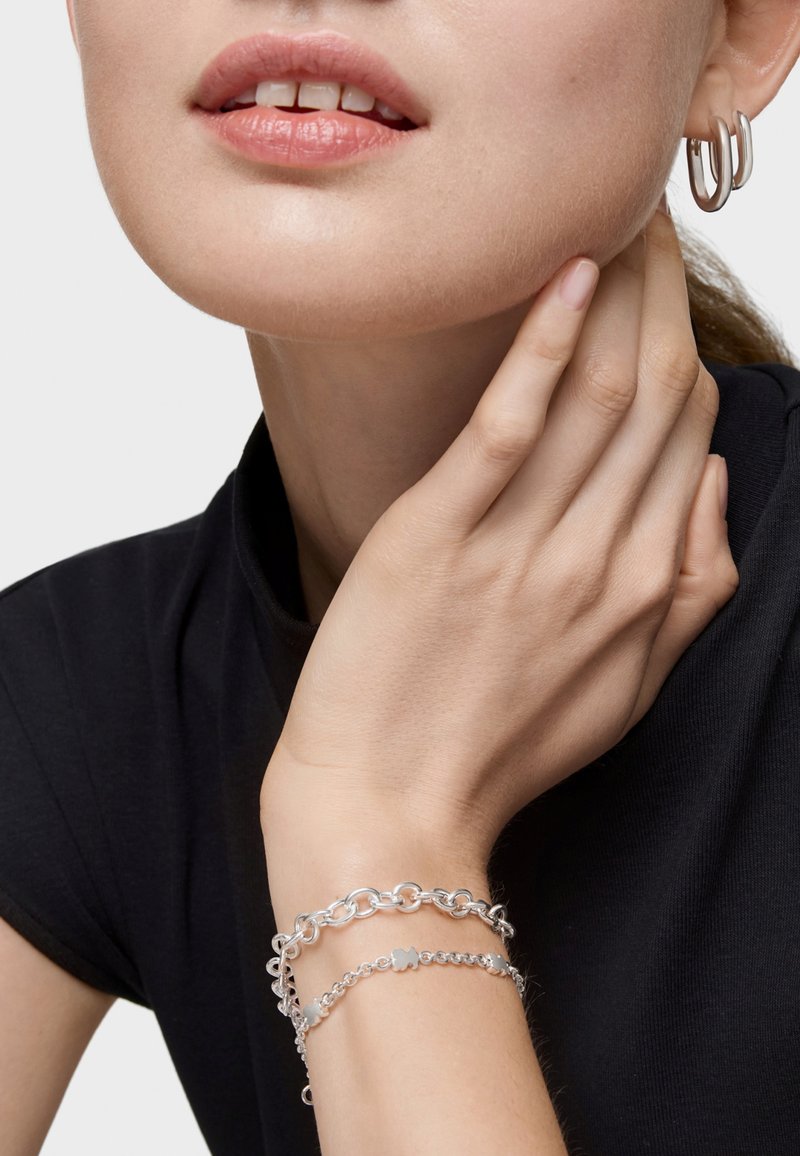 Pendientes de plata y pulseras en capas, con eslabones lisos y texturizados, llevados por una persona con una blusa de tono neutro y un maquillaje natural.