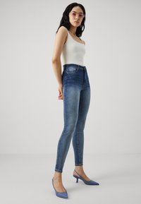 Jeans skinny de cintura alta en azul con una textura de denim suave. Combinados con una camiseta sin mangas ajustada blanca y unos tacones de slingback en azul claro.