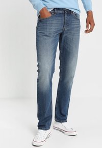 Man som bär en ljusblå tröja, medelblå raka jeans, vita sneakers, med ena handen i fickan på vit bakgrund.