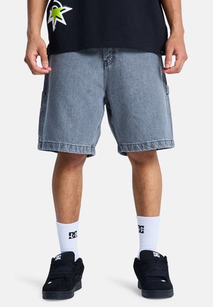 Graue Jeans-Cargoshorts mit lockerem Sitz, Seitentaschen, ausgefransten Kanten, kombiniert mit schwarzen Sneakers und weißen Socken mit Logoprint. Schwarzes T-Shirt darüber getragen.