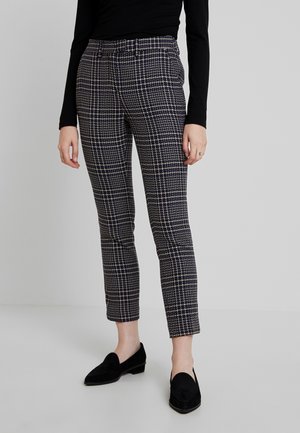 Femme portant un pantalon slim-fit à carreaux bleu marine et beige, avec des mocassins en daim noir et un haut à manches longues noir, se tenant sur un sol blanc.