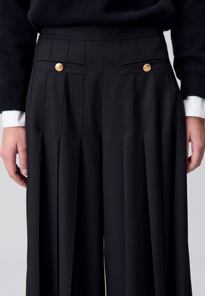 Pantaloni neri plissettati con due tasche frontali dotate di bottoni dorati e un tessuto liscio e testurizzato, che presentano un design su misura a gamba larga.