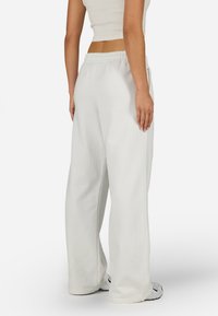 Balti platām kājām sporta bikses ar mīkstu tekstūru, regulējamu jostu un brīvu piegriezumu, kopā ar sporta apaviem.