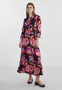 Vestido-camisa longo floral em preto, com padrões vibrantes em rosa, roxo e azul. Fecho frontal com botões, mangas compridas e corte descontraído.