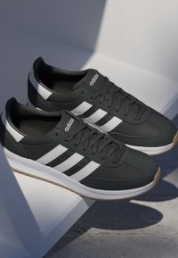 Zapatillas negras Adidas con rayas blancas y suelas de goma descansando sobre una superficie blanca inclinada bajo una iluminación suave.