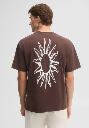 Mann trägt braunes T-Shirt mit großer weißer Sonnen-Grafik auf dem Rücken, kombiniert mit hellen Hosen, steht vor schlichtem Hintergrund.