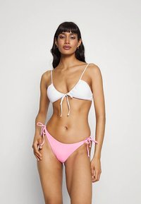 Hvit triangel-bikini topp med frontknute og tynne stropper, kombinert med rosa lavt liv bikini-bottom med sidesnorer. Glatte teksturer.