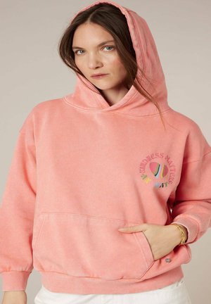 Femme portant un sweat à capuche rose avec un logo en forme de cœur coloré "La gentillesse compte", main dans la poche avant, fond clair.