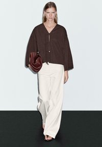 Brun, langærmet top med V-hals og snørelukning, parret med hvide, brede bukser. Modellen bærer en bordeaux, rund clutch.