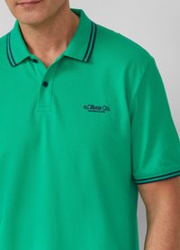 Koszulka polo w intensywnym zielonym kolorze z kołnierzykiem, dwoma guzikami i granatowymi paskami na kołnierzu. Posiada logo w ciemnym niebieskim kolorze.