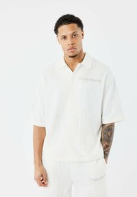 Camisa de manga corta blanca con cuello, de tejido texturizado y patrón de cactus; presenta bordado de marca en un color contrastante.