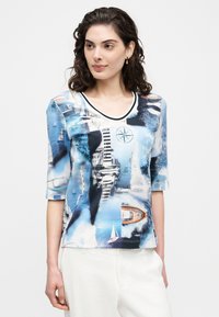 Γυναίκα με σκούρα σγουρά μαλλιά που φοράει μπλε τοπ με ναυτικό σχέδιο, με βάρκες, φάρους και πυξίδα, συνδυασμένο με λευκό παντελόνι.