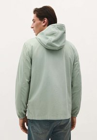 Lys grøn hættejakke med elastiske manchetter, en afslappet pasform og en glat tekstur, parret med blå jeans. Bagfra set.