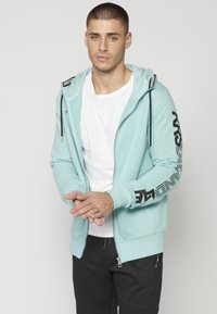 Sudadera con cremallera de color azul claro con gráficos de texto negro en las mangas, bolsillos frontales y cordones. Lleva puesta una camiseta blanca debajo.