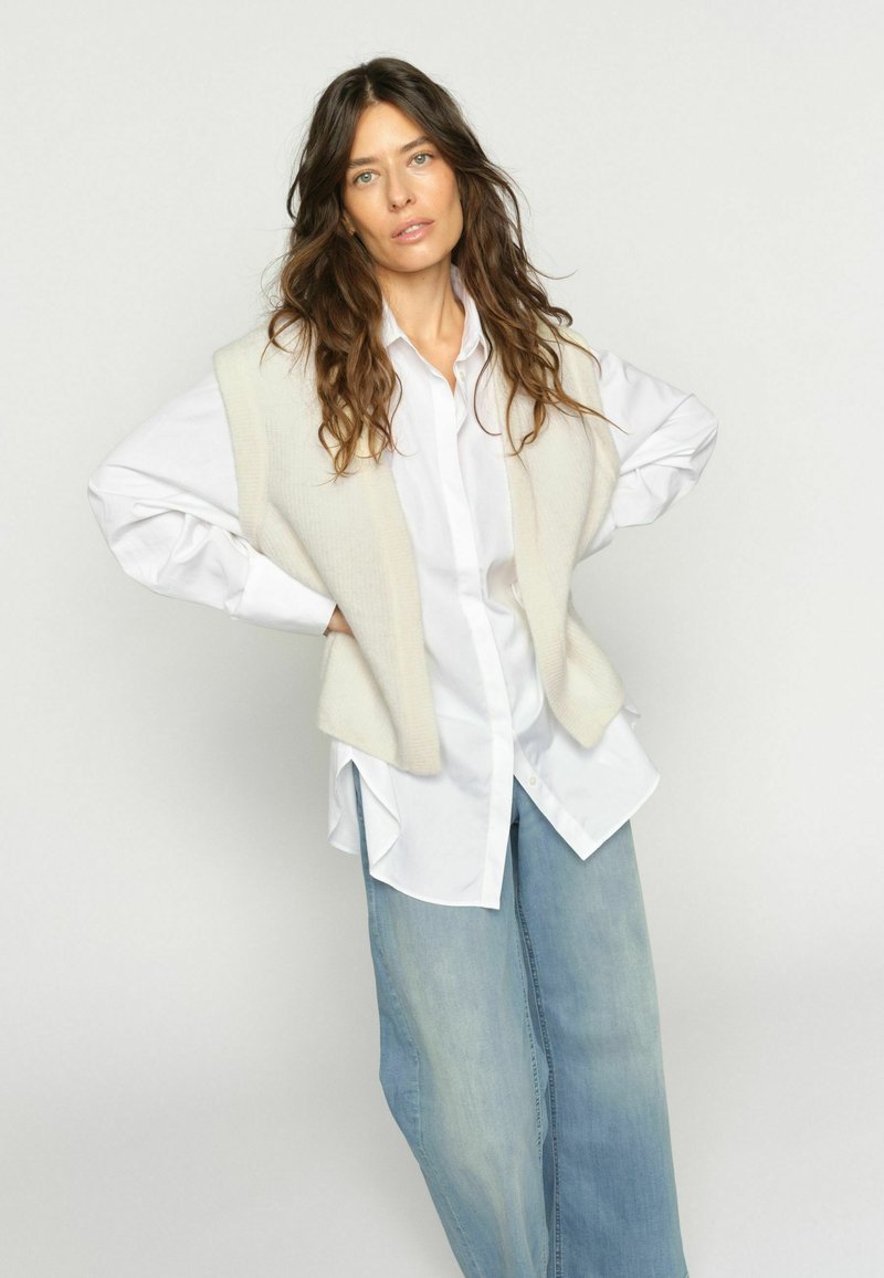 Femeie cu păr lung și ondulat, purtând o cămașă albă cu nasturi, un vestă tricotată crem și blugi largi, albastru deschis, pe un fundal simplu.