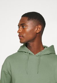Hooded sweatshirt in muted green fabric with a smooth texture; features drawstrings and metal hardware at the neckline.