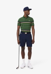 Polo a righe verdi, pantaloni corti blu e scarpe bianche. Tiene un bastone da golf. Accessori includono un berretto blu e una cintura nera con fibbia argentata.