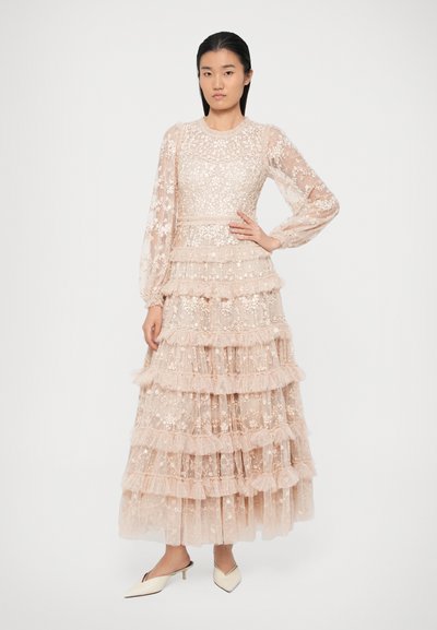 Ilga švelni rožinė nėrinių suknelė su sluoksniuotais volanais, permatomomis ilgomis rankovėmis, gėlių siuvinėjimu ir prigludusia krūtine. Suknelė derinama su baltais smailiais aukštakulniais.