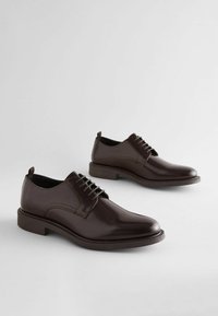 Zapatos de vestir de cuero marrón con un acabado pulido, punta redonda, diseño con cordones y una suela contrastante. Incluye una pequeña lengüeta en el talón.